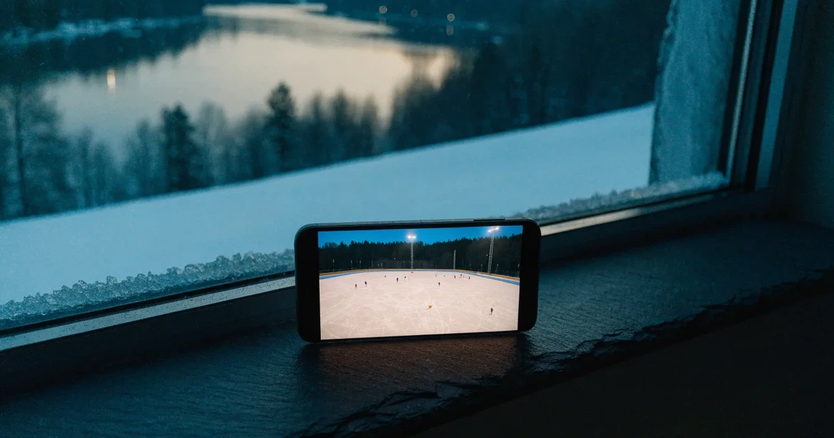 Eine Eisfläche unter Flutlicht in der Winterdämmerung, aus grosser Distanz von einem verschneiten Hang aus gesehen.