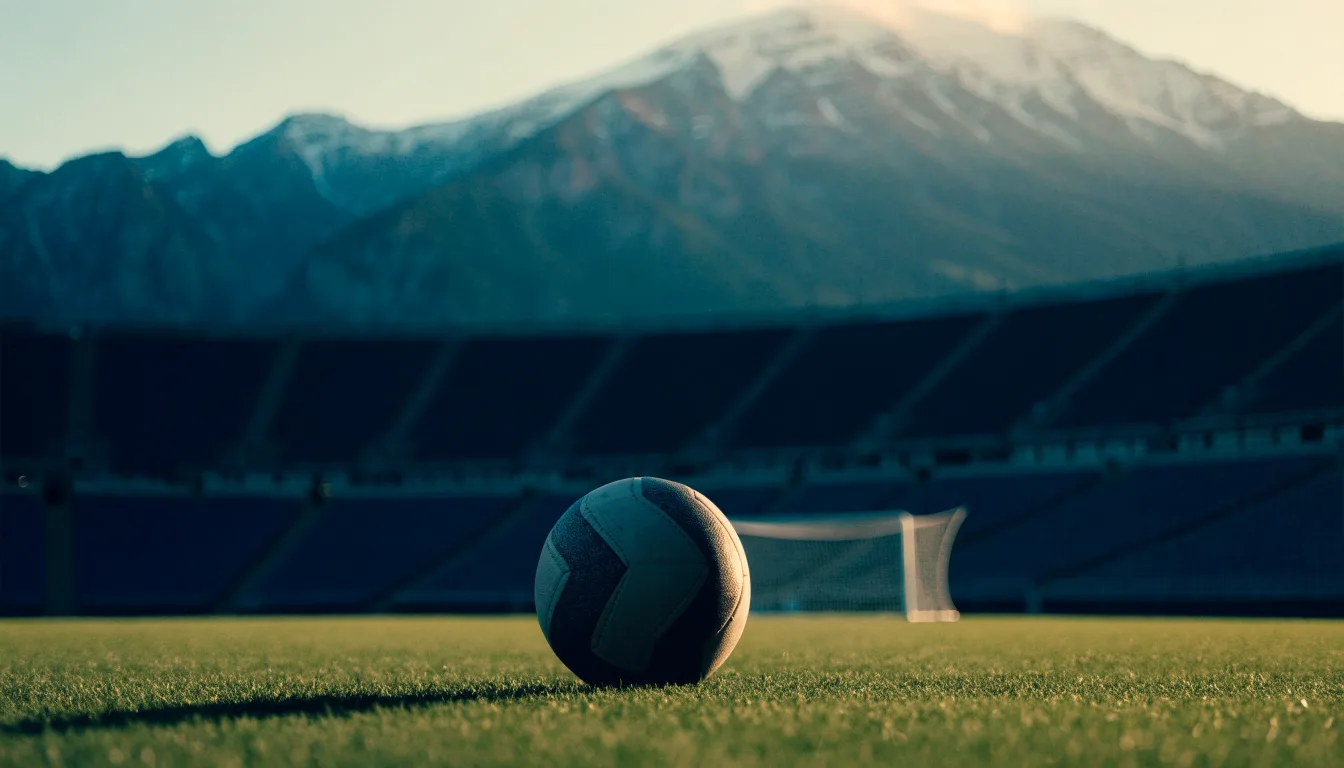 Ein Fussball liegt auf dem Rasen eines leeren Stadions am frühen Abend, dahinter erheben sich schneebedeckte Berge unter klarem Himmel.