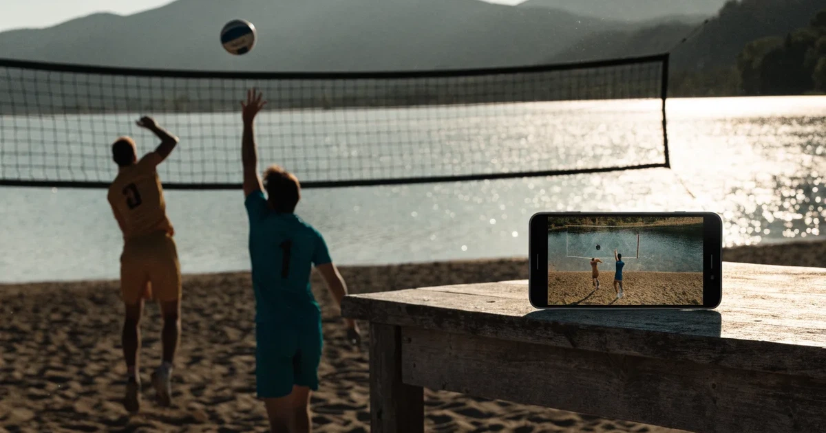 Ein Volleyball liegt am Fuss des Netzes auf Sand, im Hintergrund glitzert ein See und ein Bergzug steht im Dunst.