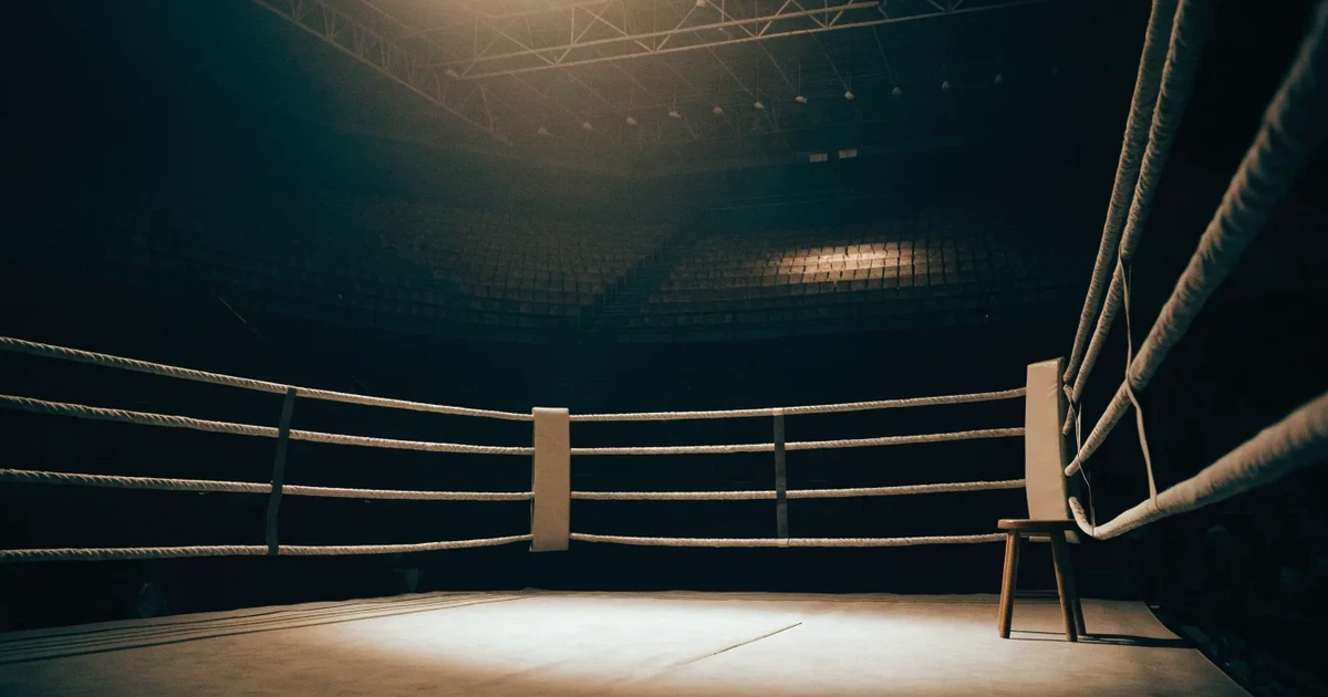 Ein leerer Boxring mit gespannten Seilen unter hellem Hallenlicht, dahinter ansteigende Zuschauerränge in einer grossen dunklen Halle