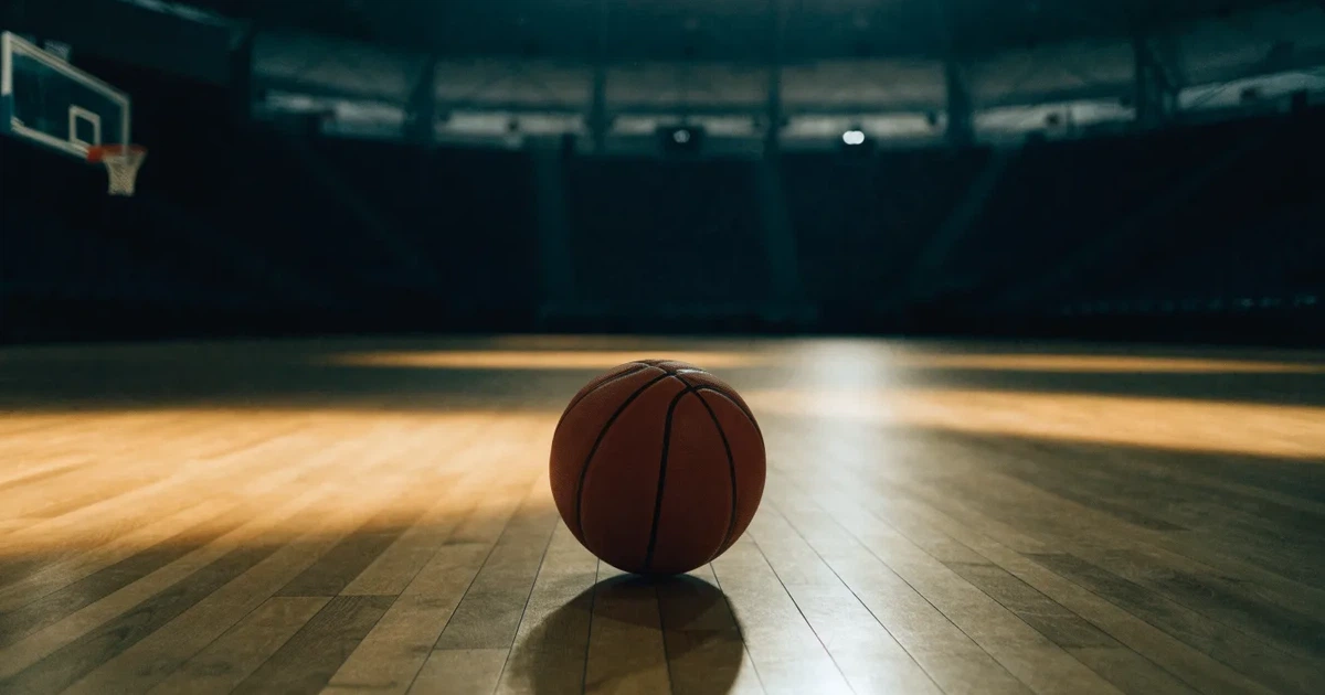 Ein Basketball liegt auf dem hellen Parkett einer leeren Halle, im Hintergrund steigen die Tribünenreihen im Halbdunkel an.