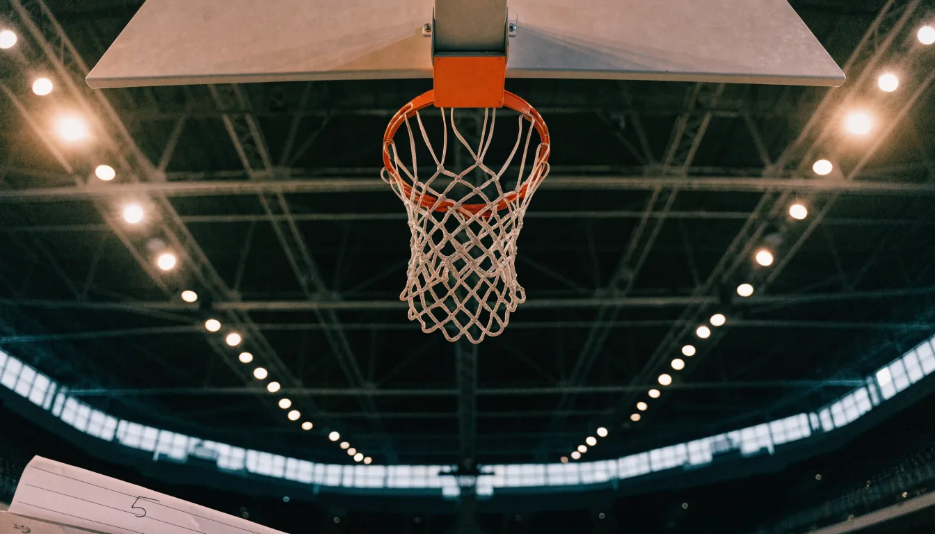 Ein Basketballkorb von unten gesehen, darüber eine Hallendecke mit Scheinwerfern und Stahlträgern.