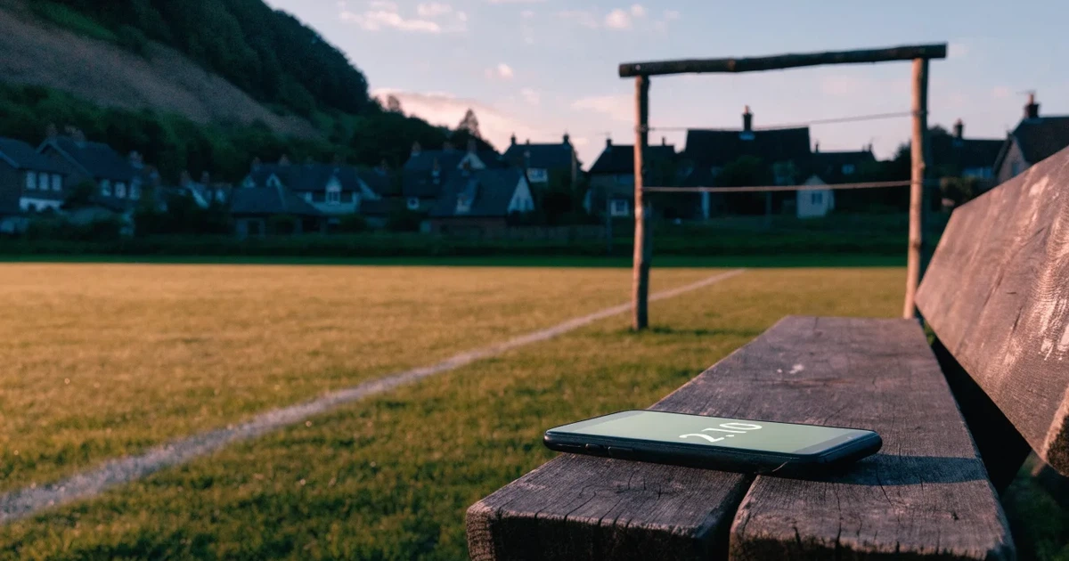 Ein kleiner Fussballplatz mit einfacher Holztribüne und Rasenstreifen am Rand einer Ortschaft, im Hintergrund ein bewaldeter Hügel im Abendlicht.