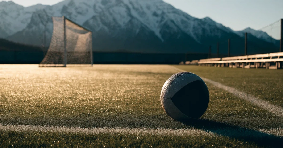 Ein Fussball liegt am frühen Morgen auf dem Rasen eines leeren Fussballfeldes, dahinter eine kleine Tribüne und schneebedeckte Berge.