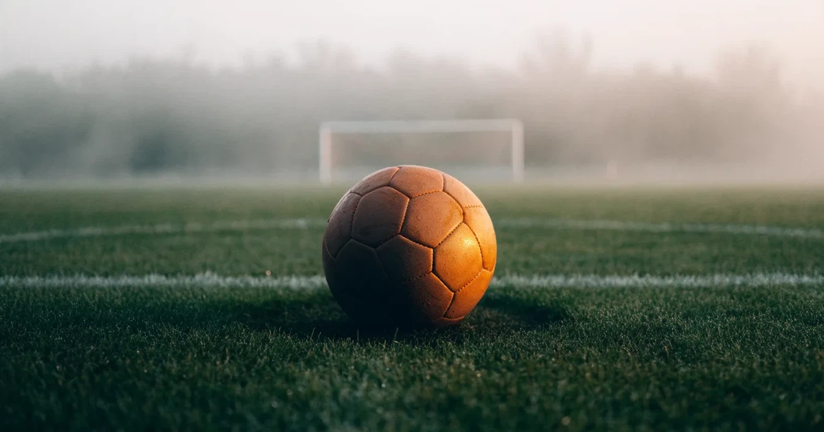 Ein Fussball liegt auf dem Anstosspunkt eines leeren Rasenplatzes im dichten Morgennebel.